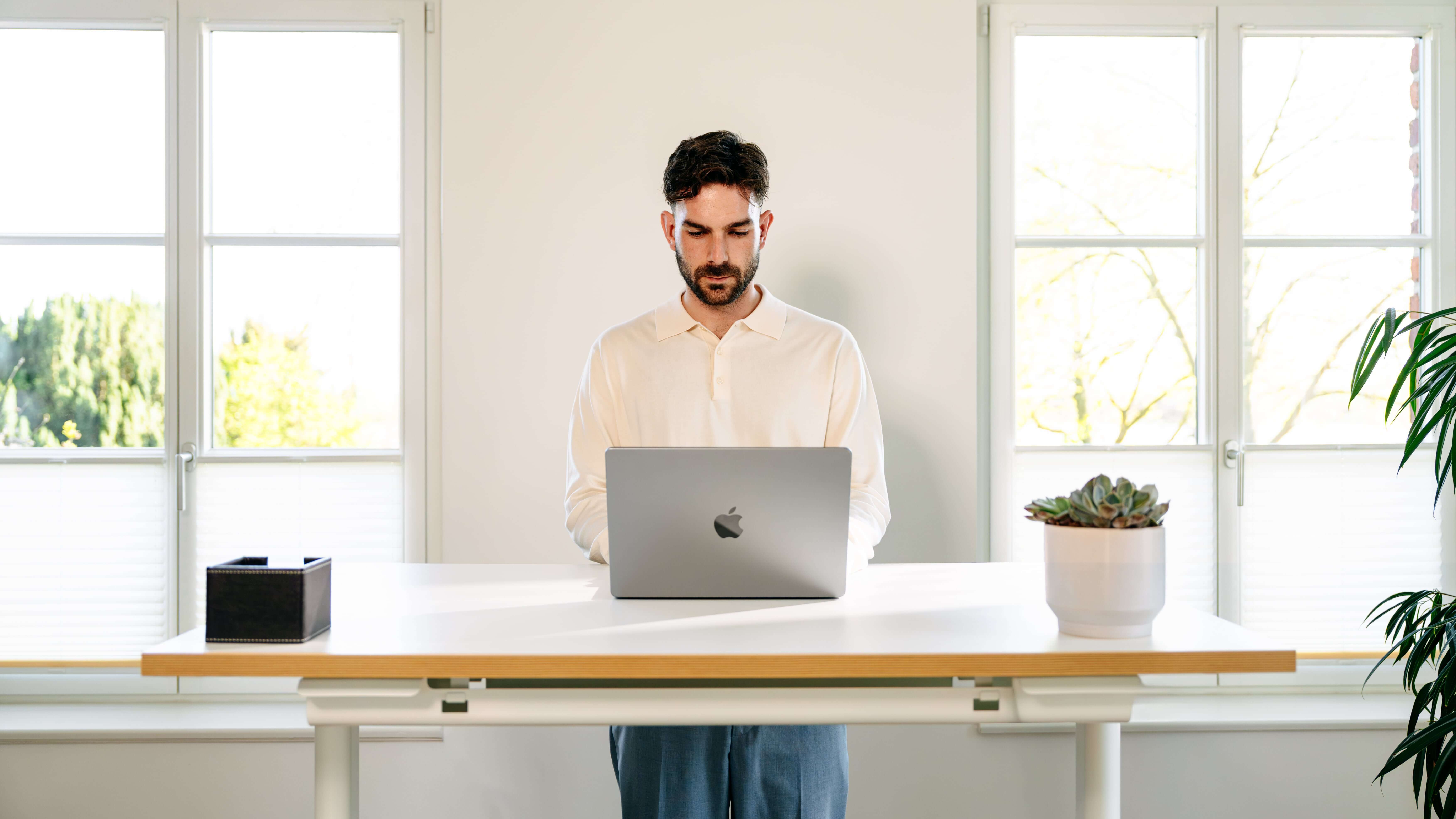 Un homme travaille debout devant un bureau réglable en hauteur, équipé d'un ordinateur portable, sur fond clair et entouré de plantes, dans un bureau moderne.