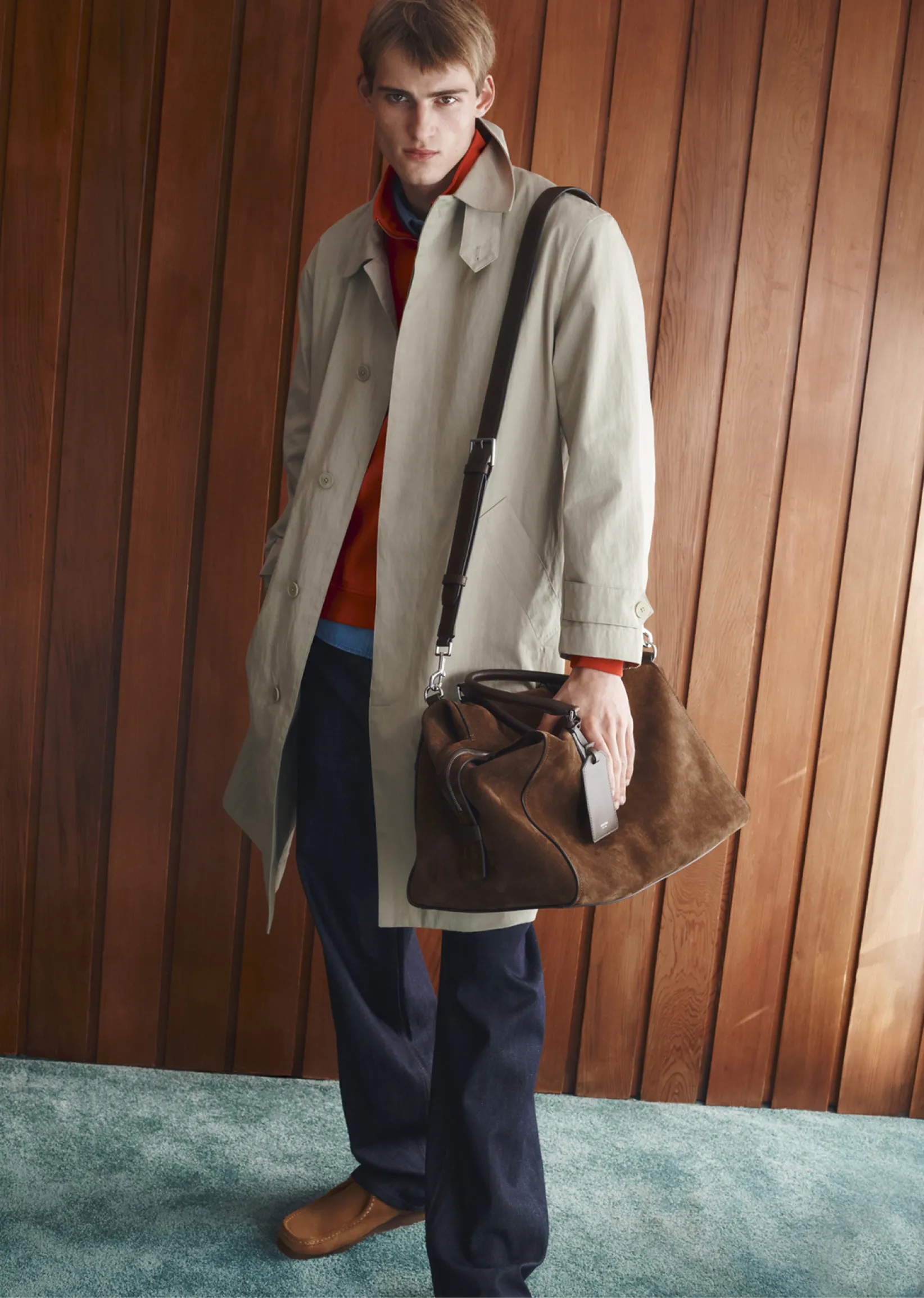A model in a trench coat stands against a wooden wall, holding a bag.