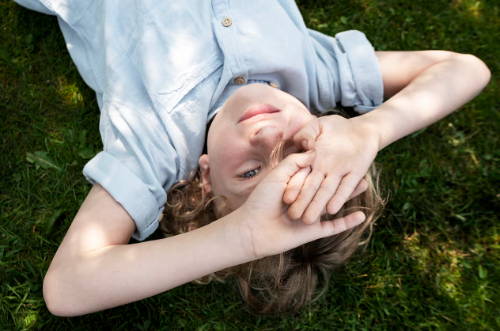 Ein glückliches Kind entspannt sich auf dem Rasen und schützt mit den Händen die Augen vor der Sonne. Die Allergie-Immuntherapie gegen Allergien kann dazu beitragen, die Symptome der Gräserallergie zu reduzieren.