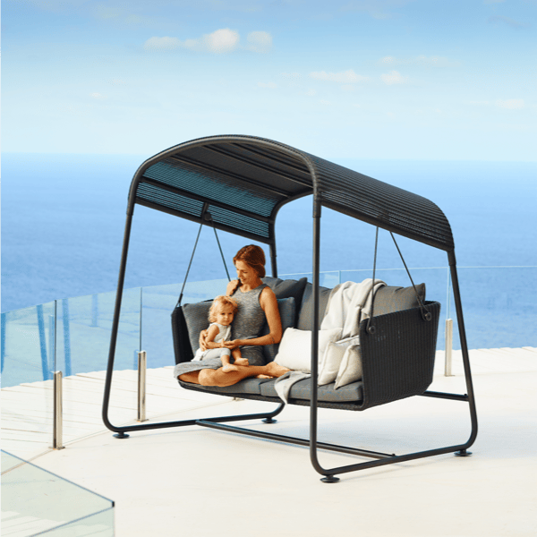 The Cave Outdoor Swing Sofa with a sleek black metal frame, gray woven seating, and plush cushions, set against a scenic ocean view. Ideal for relaxed, playful outdoor lounging and romantic seating.