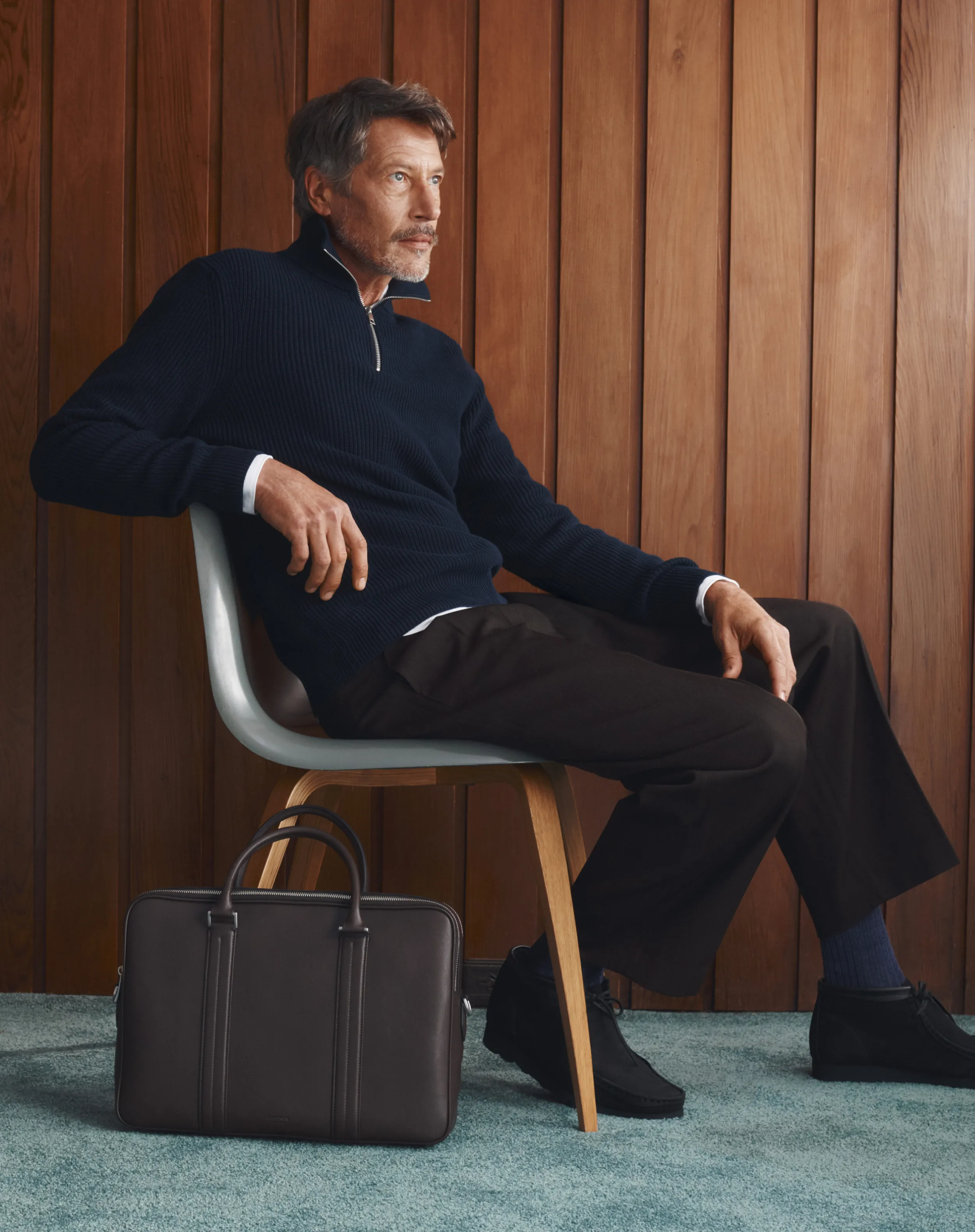 Man sitting on a chair, wearing a sweater, with a bag beside him.