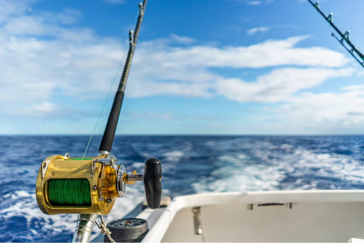 Close-up of a gold offshore fishing reel with green fishing line mounted on a boat rod holder, overlooking open ocean water and boat wake under a bright blue sky.