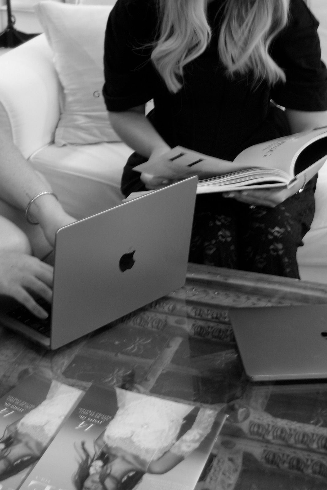 Two people working on laptops with magazines on a table.