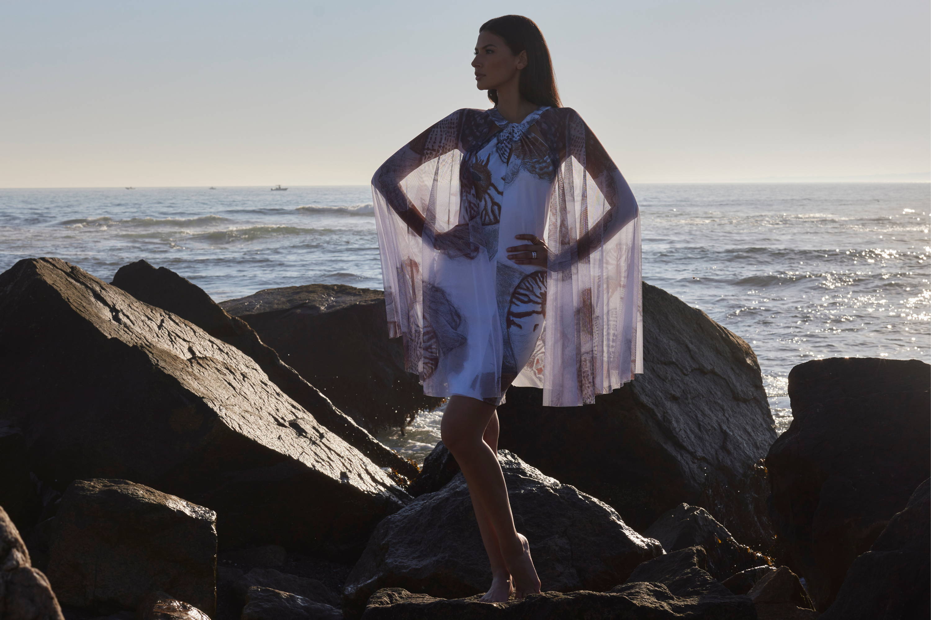 Woman wearing stretch knit short dress with mesh cape attached to the collar on the rocks by the ocean by Ala von Auersperg for resort 2024