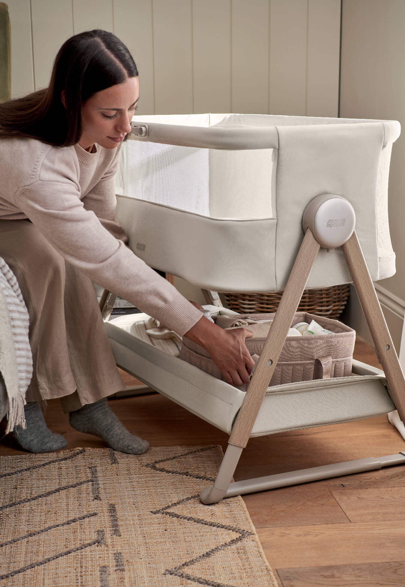Woman adjusting a baby bassinet with storage underneath in a cozy room.