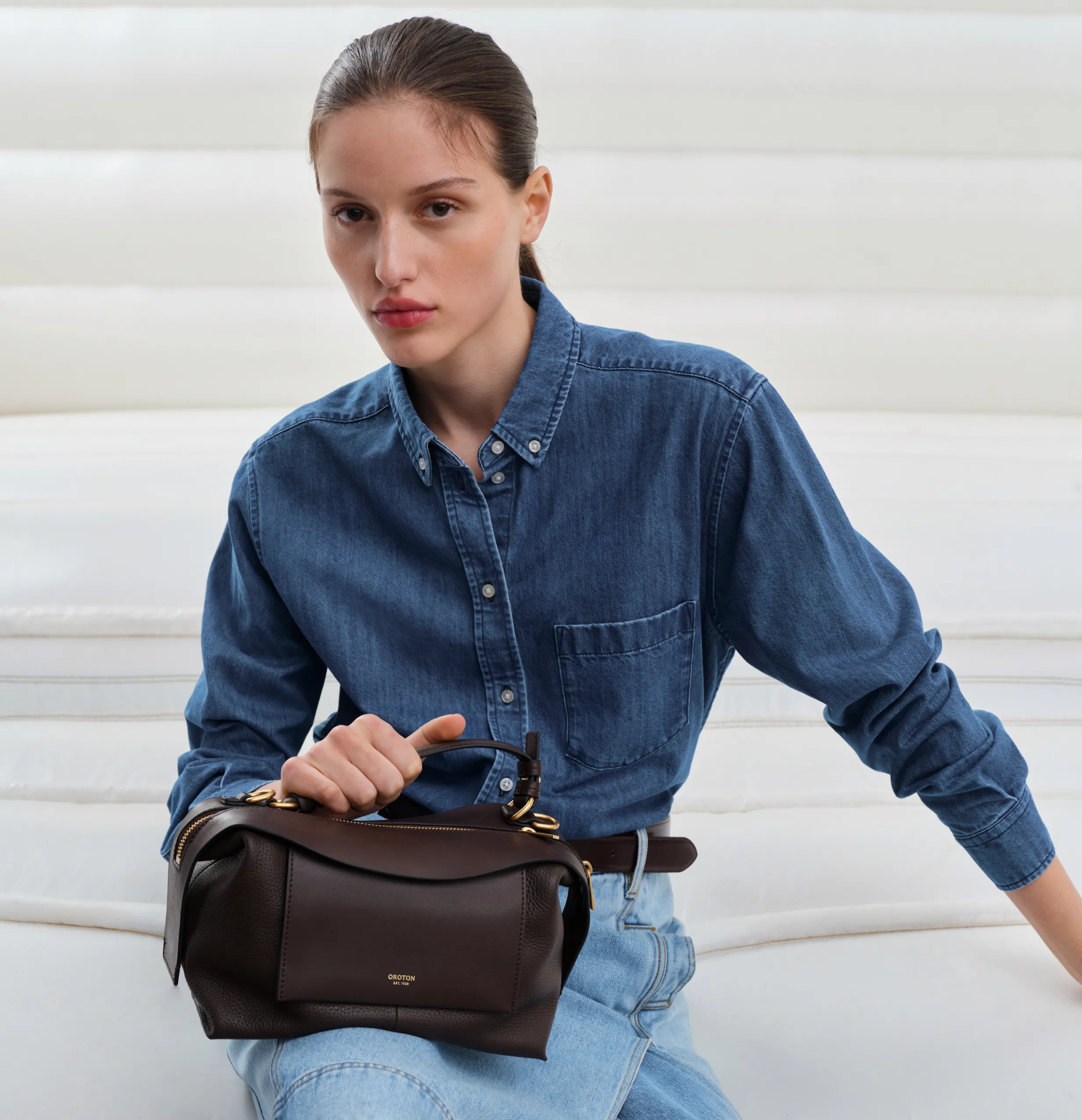 Woman in denim shirt holding a brown handbag, seated on a white couch.