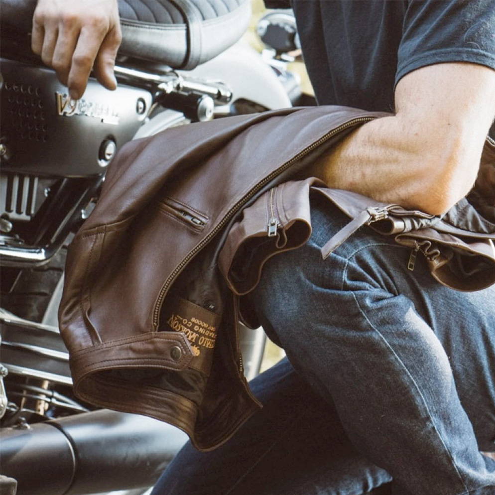man sitting next to motorcycle holding a brown motorcycle leather jacket