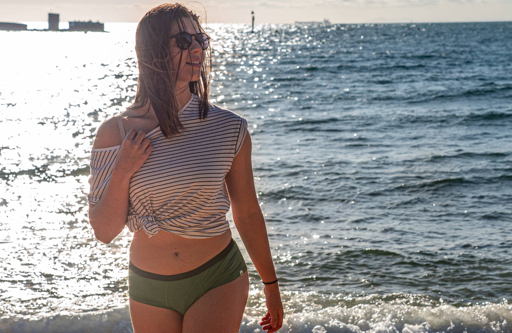 A woman in green underwear, a striped shirt, and sunglasses walking away from the ocean.