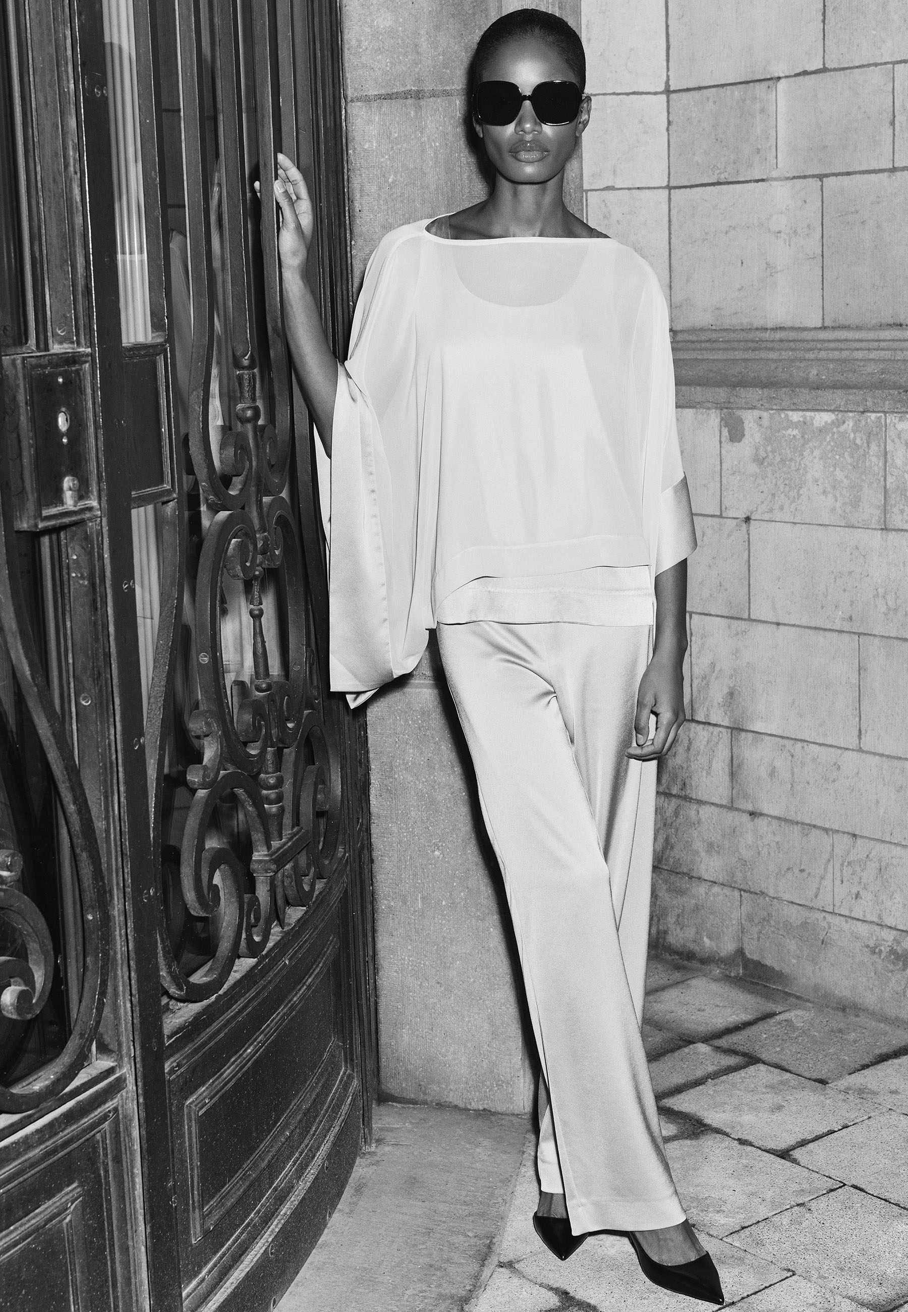 Woman in stylish outfit posing near ornate door, wearing sunglasses. Black and white photo.