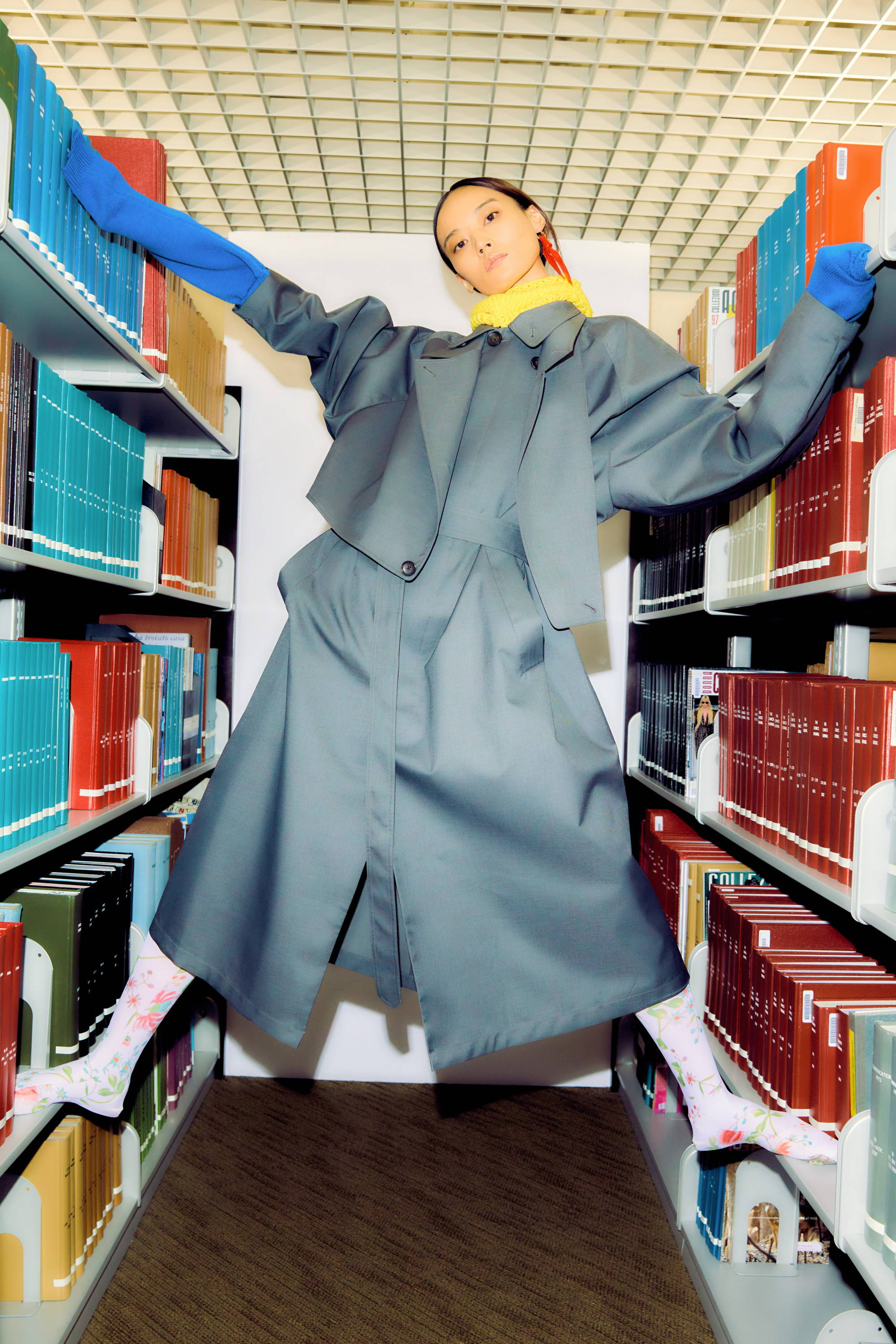 Person in a gray coat posing between colorful bookshelves in a library.