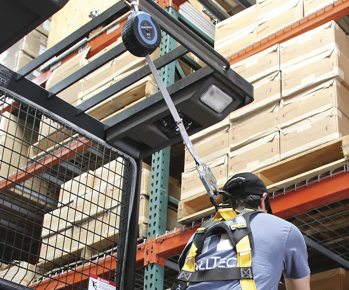 Warehouse worker grabbing boxes for an order connected to a compact overhead self-retracting lifeline anchored to his cherry picker