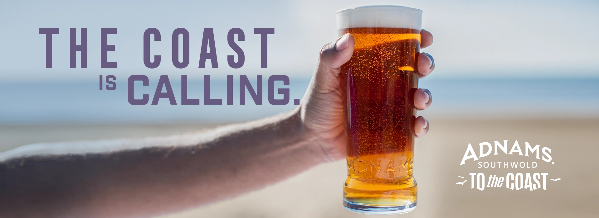 A hand holding a pint of beer with a beach background.
