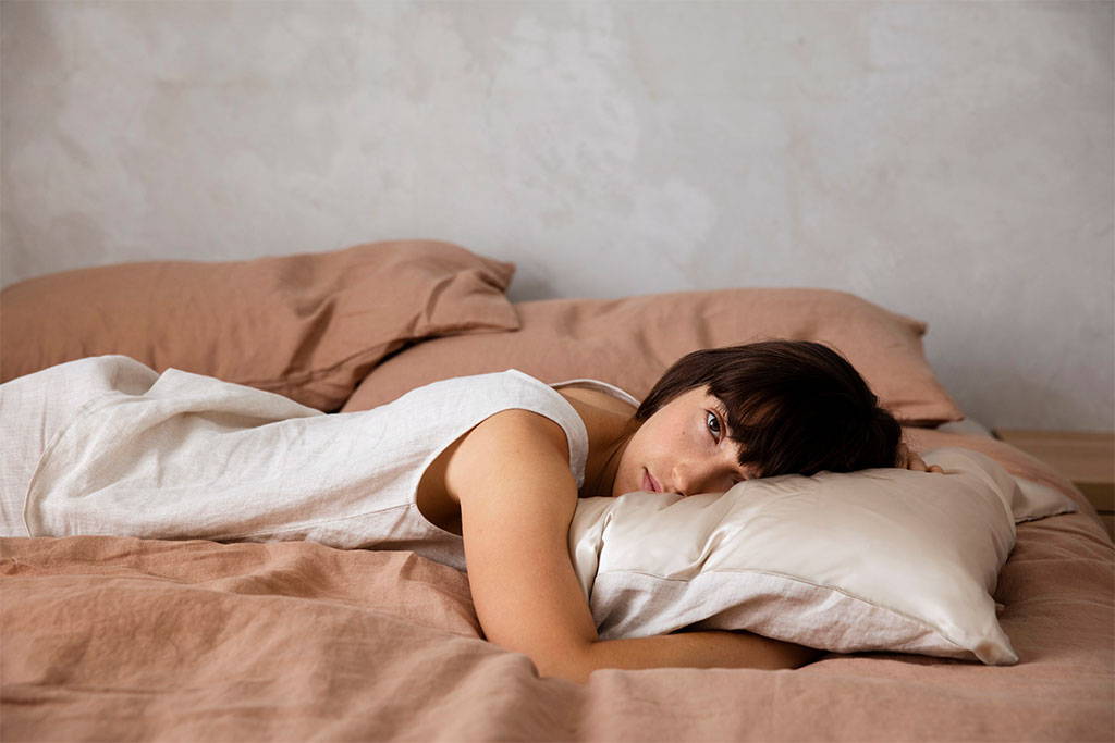 Model in a beige singlet and shorts lying on a bed dressed in the Linen Duvet Cover Set in Fawn. Model is resting her head on the Silk Linen Flip Pillowcase in Natural.