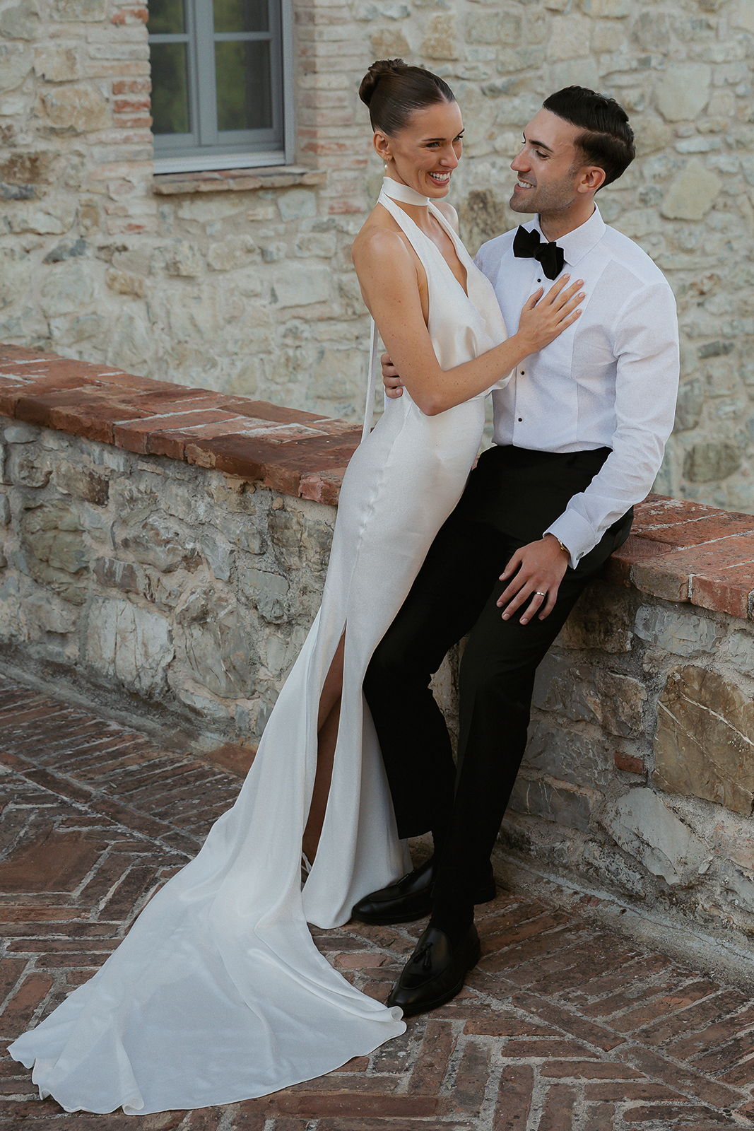 Couple in elegant attire poses together near a stone wall.