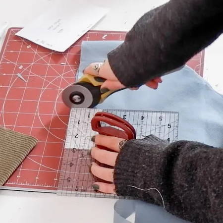 hand holding a rotary cutter, cutting blue fabric with a ruler on a rotating cutting mat