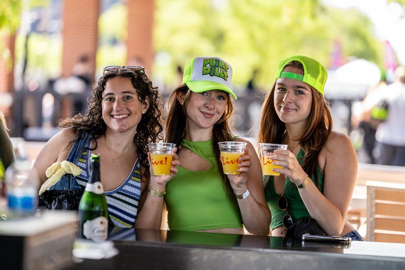 Three friends in neon trucker hats and VIP wristbands raising mimosas at the VIP Brunch Party, returning to 2100 Social at The Big Dill® Philadelphia pickle party on National Pickle Day