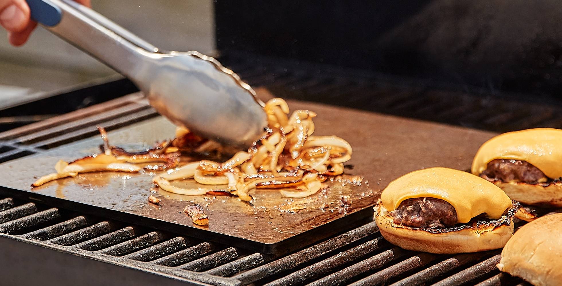 A Misen Oven Steel on an outdoor grill caramelizes thin onion slices while cheeseburgers in buns finish cooking nearby.
