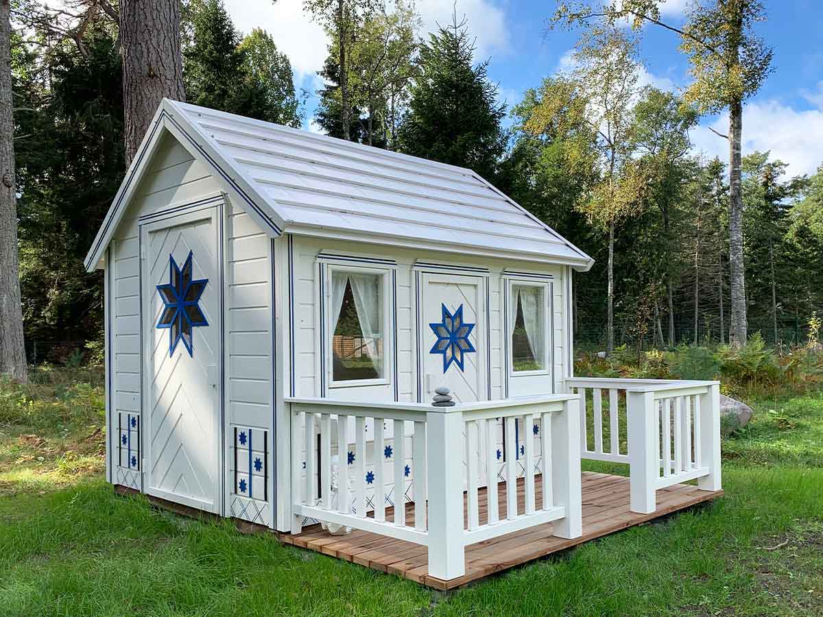 White wooden playhouse with a porch in front of forest on green grass by WholeWoodPlayhouses