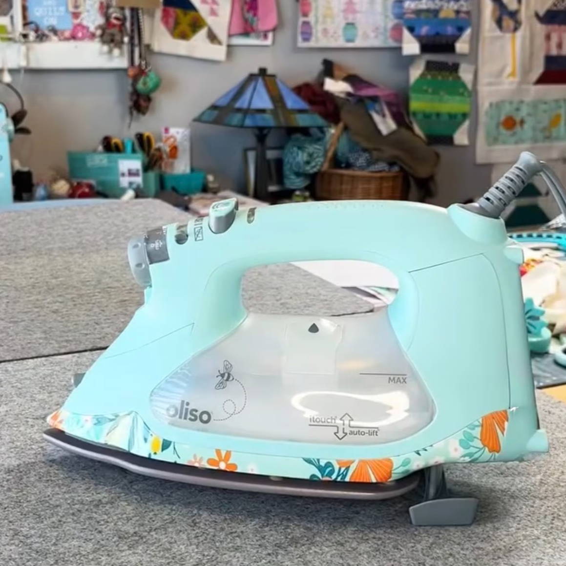 A light blue iron on a gray surface with colorful decor in the background.