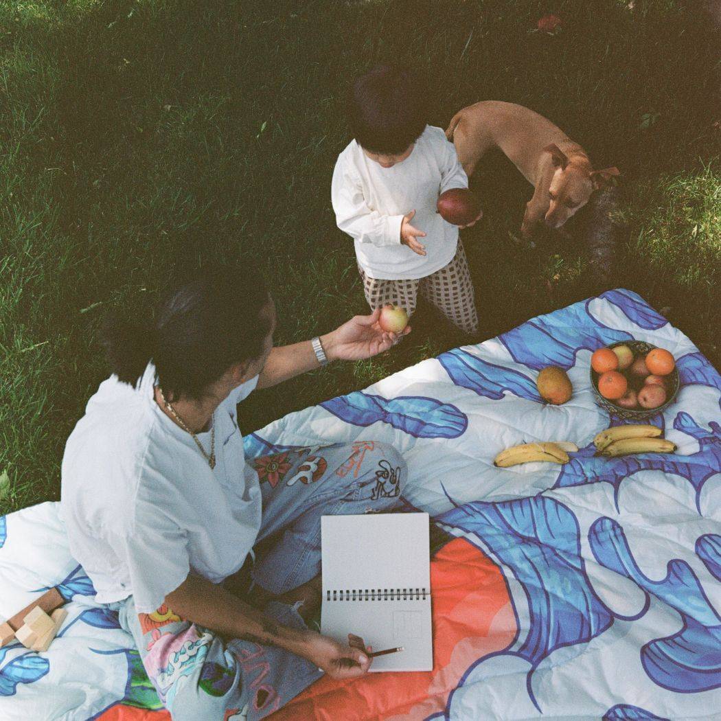 Artist and father Aaron Kai sketches on a Rumpl blanket outside as his son and dog approach him with snacks.