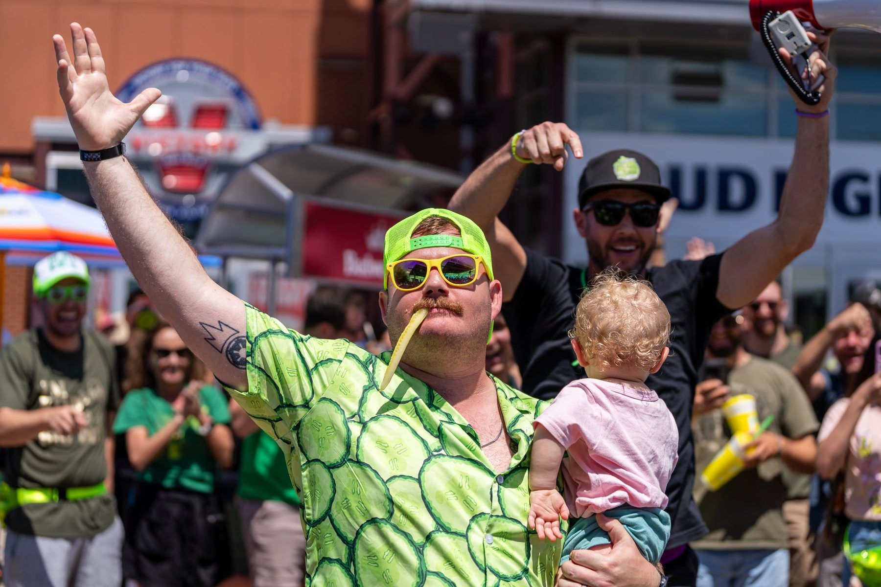 Guest in pickle-print shirt with pickle in mouth holding a toddler at The Big Dill Baltimore 2025.