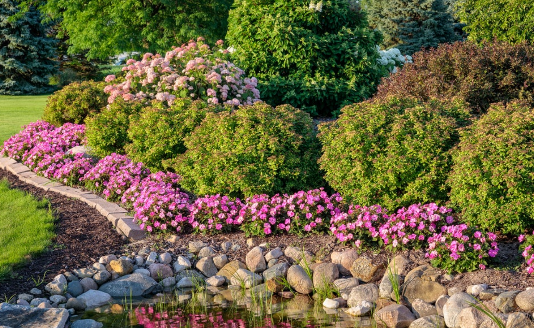 Garden and rock pond filled with SunPatiens® Compact NeoInvincibelle® Spirit II, SunPatiens® Compact Neon Pink, Double Play® Gold n Pink, Supertunia Mini Vista® Hot Pink, SunPatiens® Compact Tropical Rose, Graceful Grasses® Vertigo® 