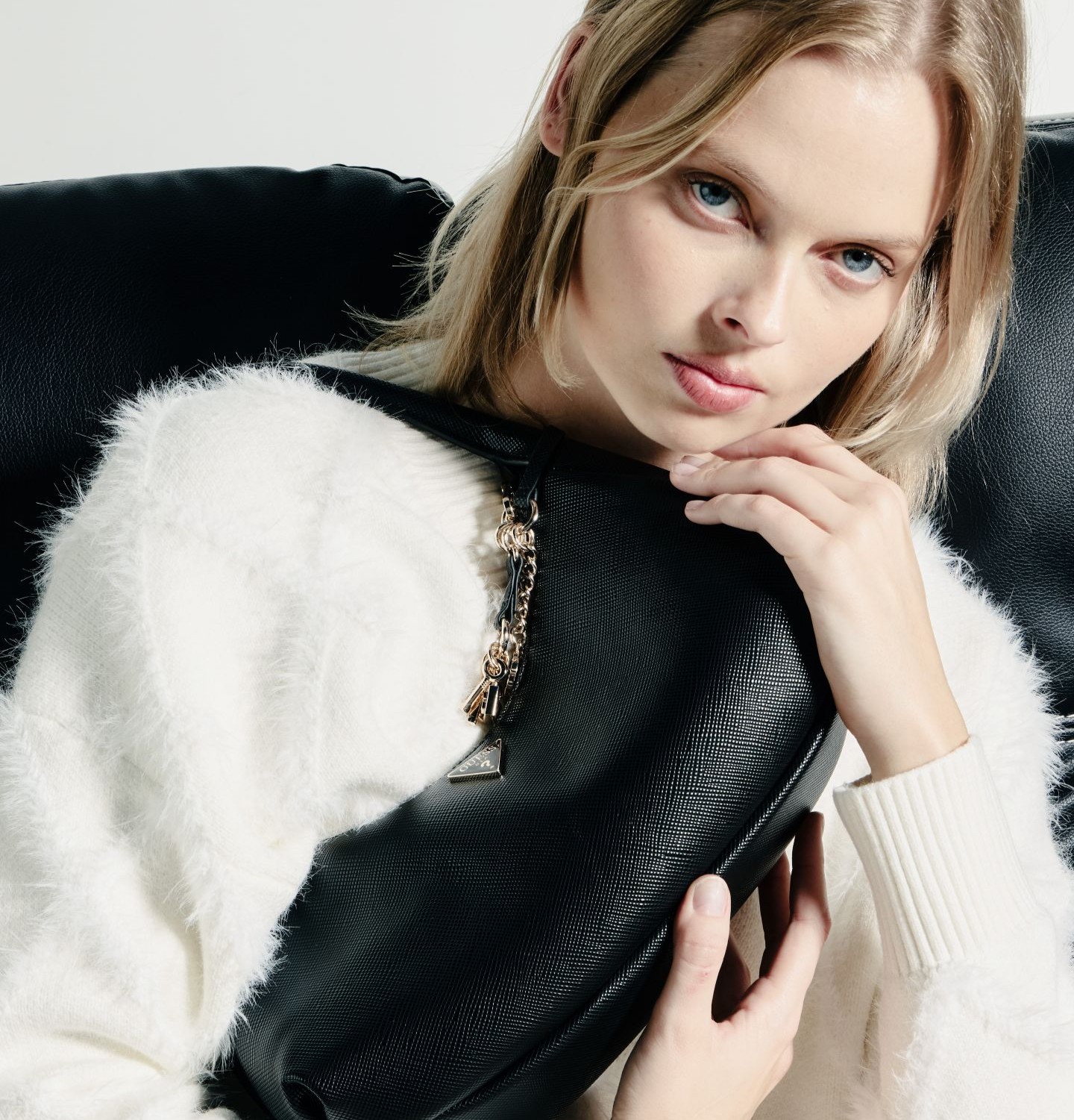 Woman in a white sweater holding a black bag, seated on a black chair.