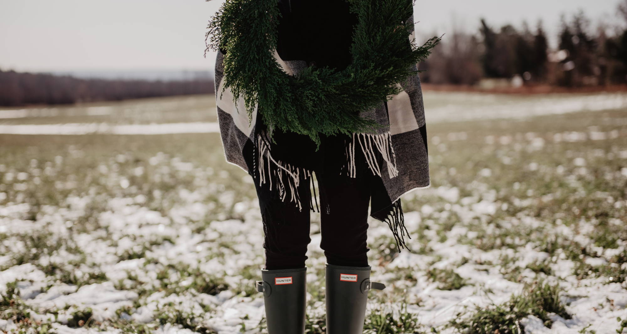 Woman wearing layers of tall black boots, skinny jeans, a cardigan, and a scarf while holding a wreath in the snow.