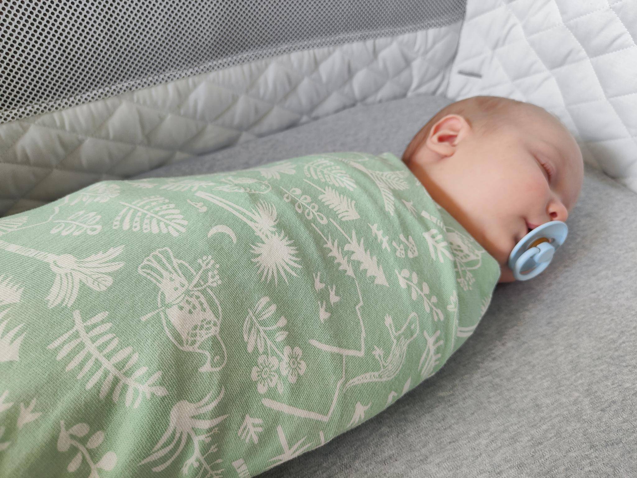 Newborn baby asleep in bassinet - swaddled in a Woolbabe Moss Wilderness swaddle blanket