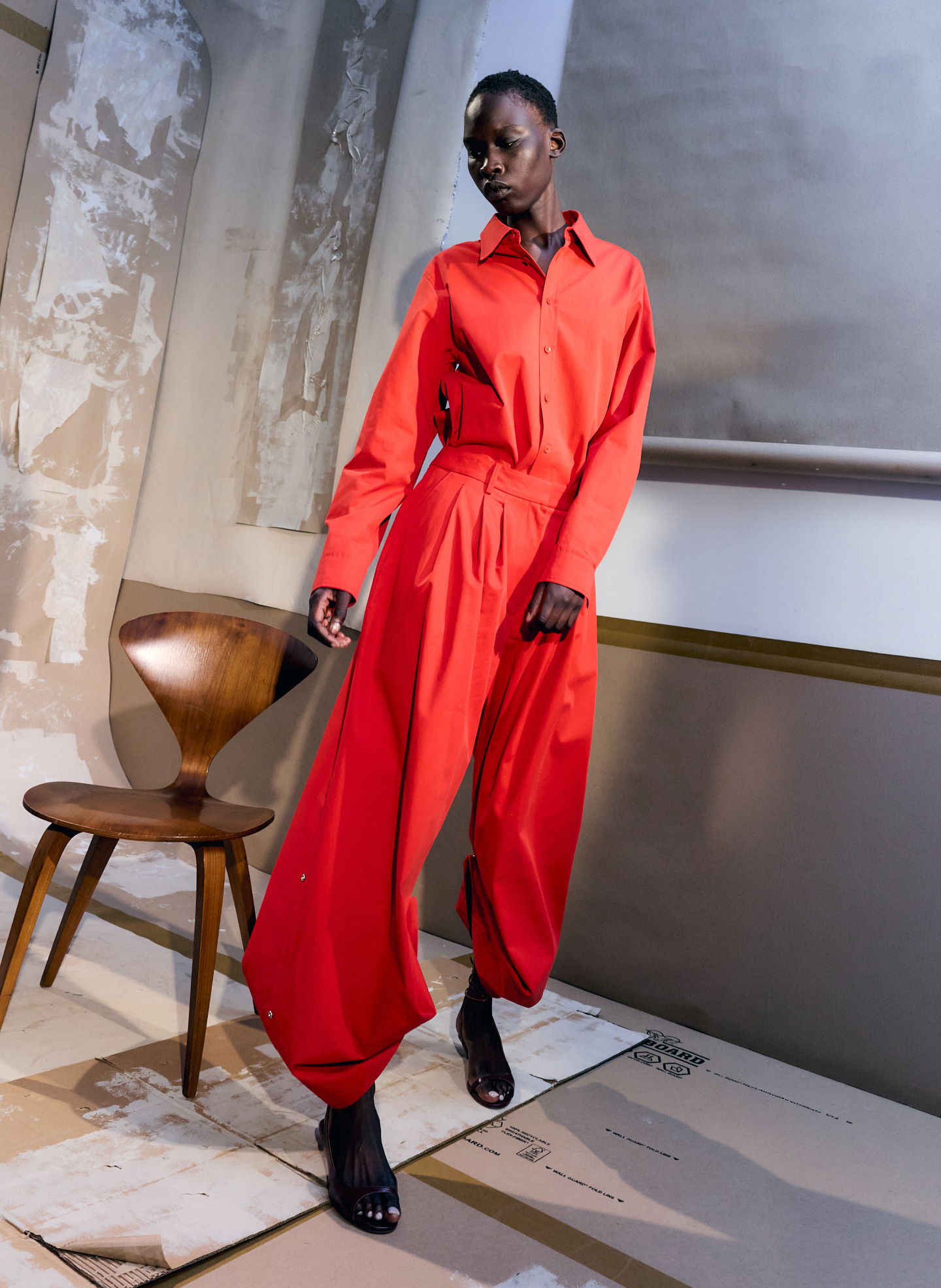 Model wearing a red outfit, standing next to a wooden chair in an artistic setting.