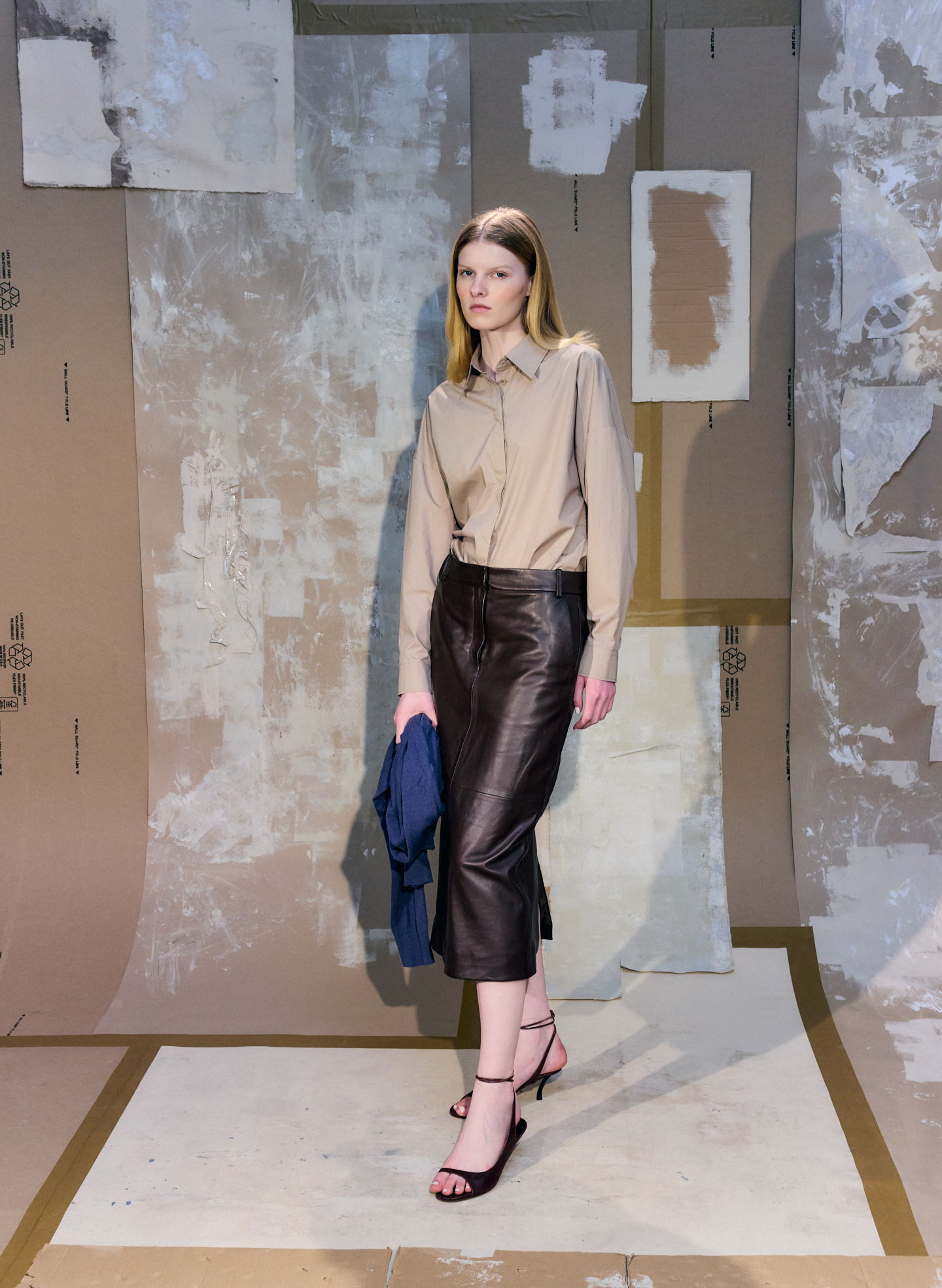 Model walking on runway, wearing a beige shirt and black skirt.