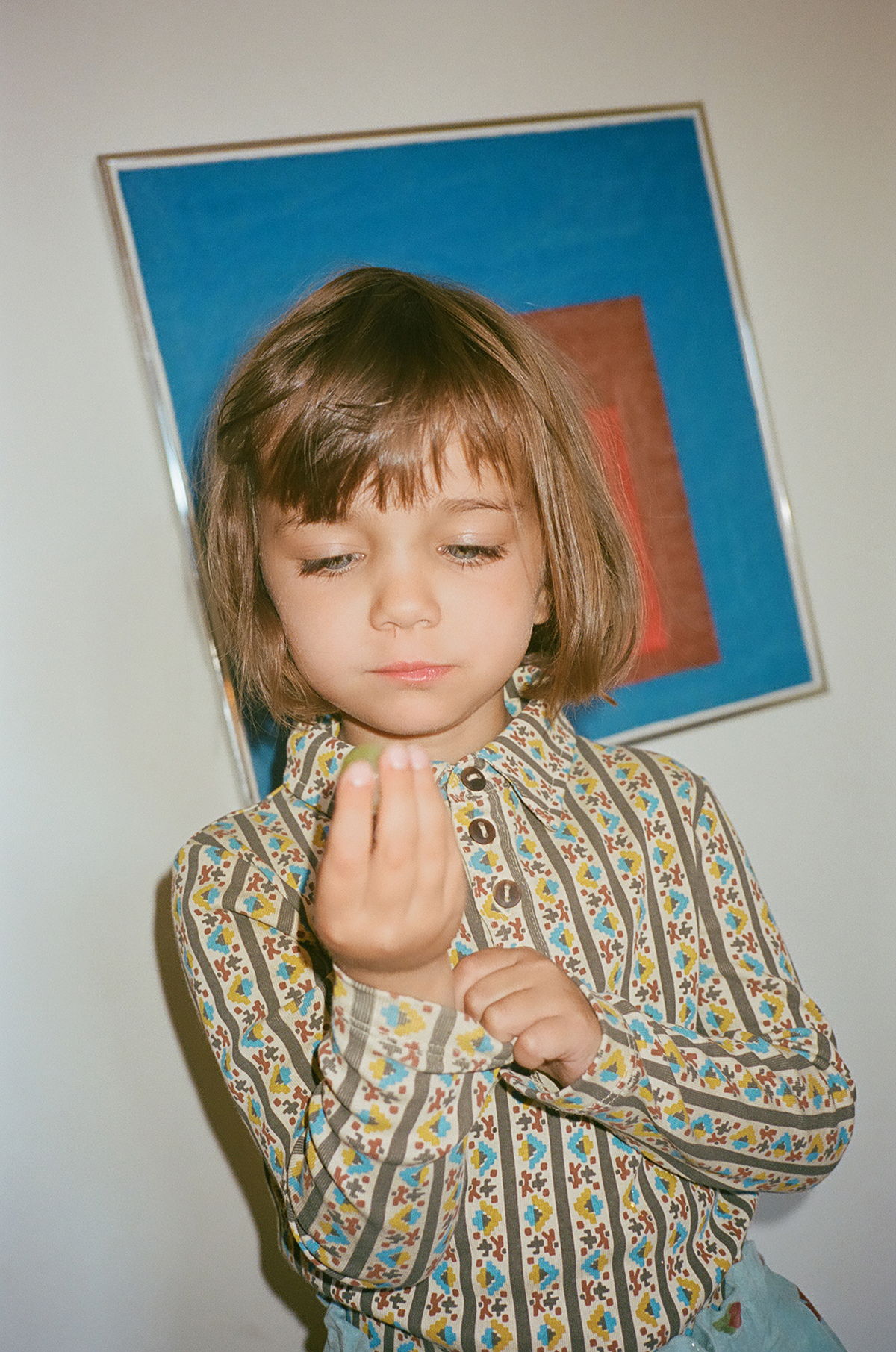 Child holding something, wearing patterned shirt, with abstract art in background.