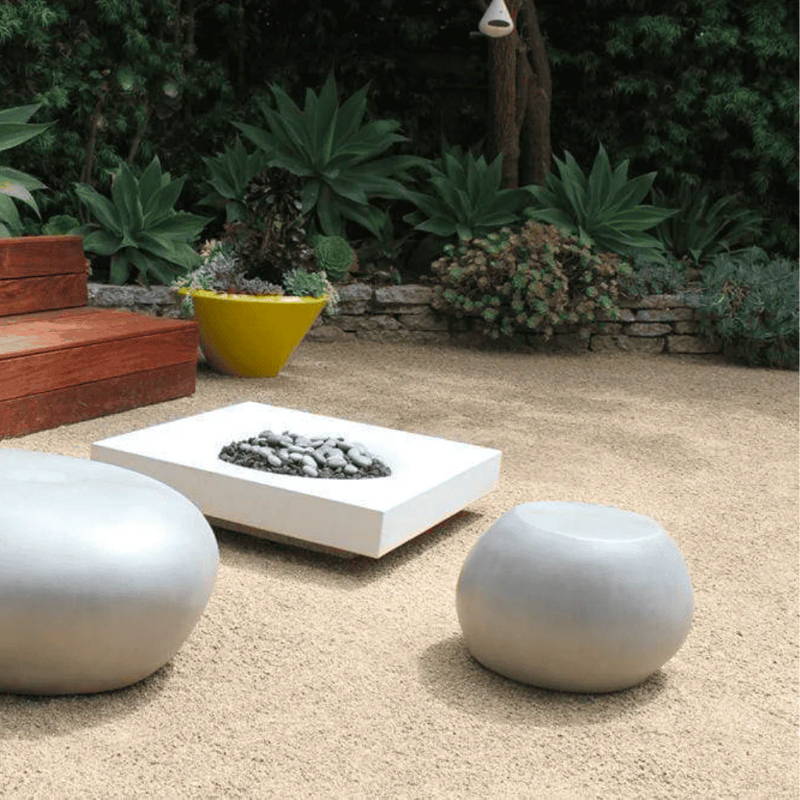 An outdoor pebble garden table sits beside a low white fire pit on a gravel patio with lush plants growing in the background.