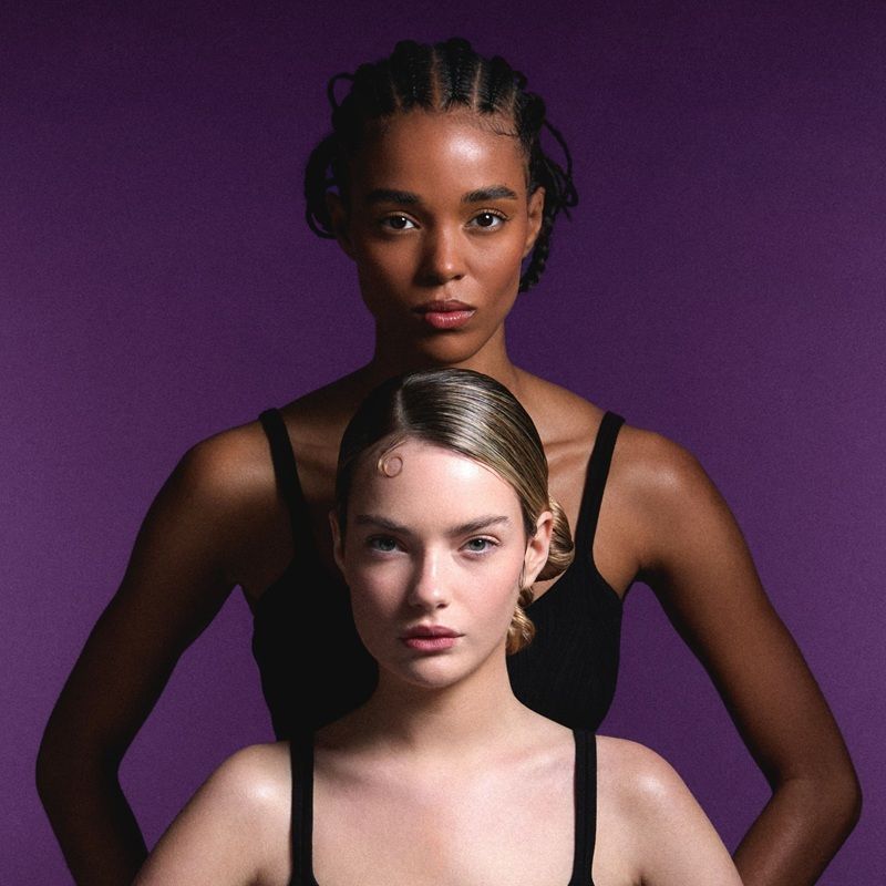 Two women with different skin tones, posing against a purple background.