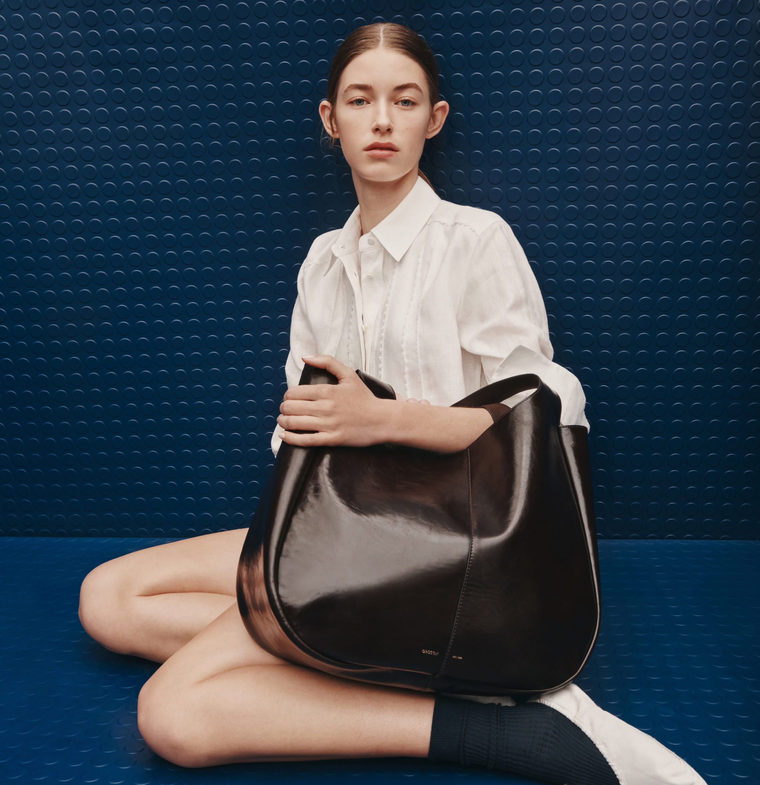 Model sitting with a black handbag against a blue background.