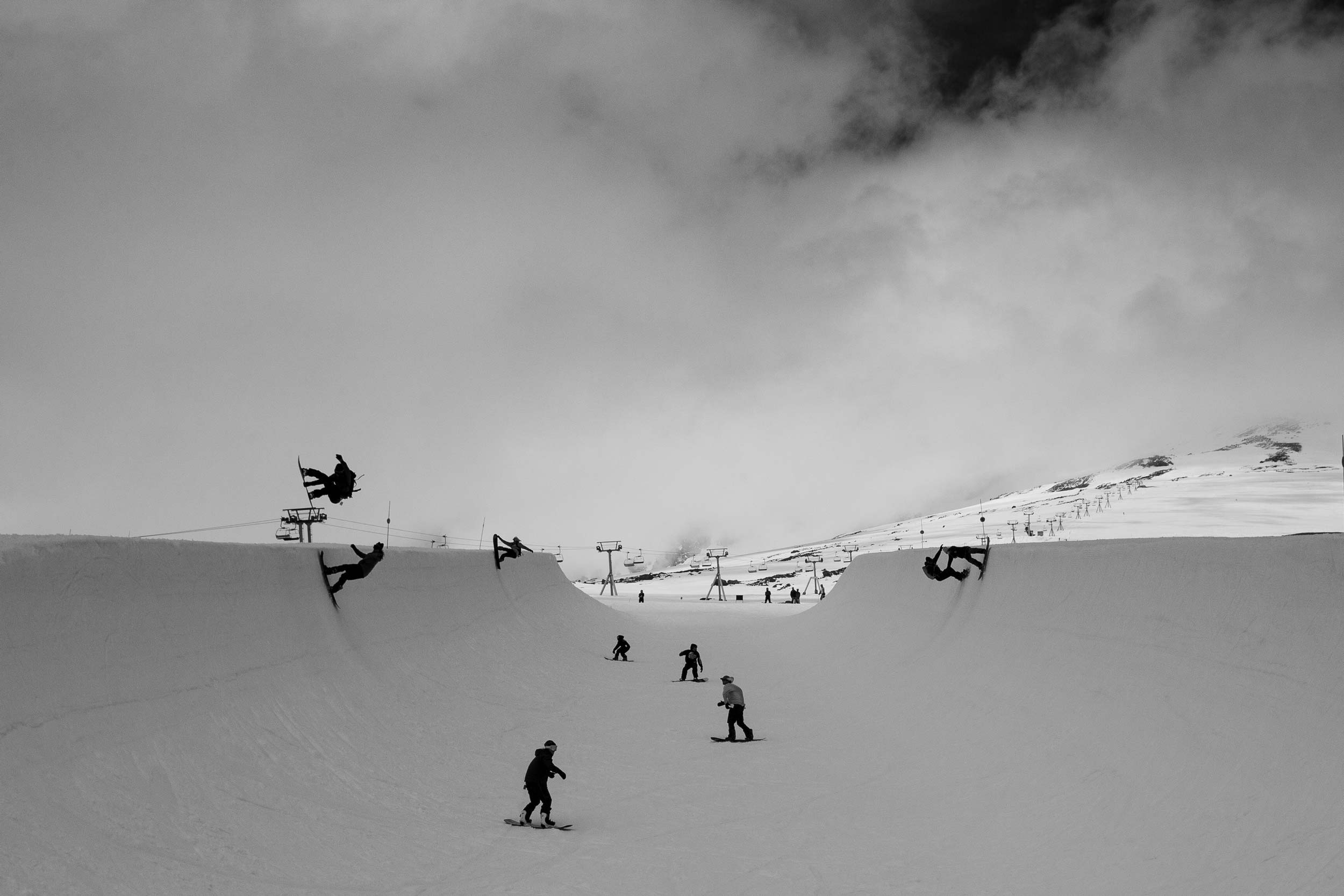 Snowboarders performing tricks in a snowy landscape, captured in black and white.