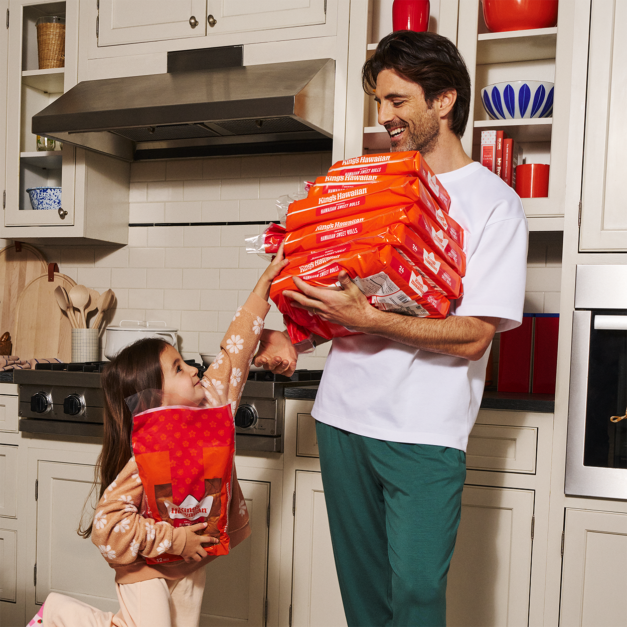 Adult man carrying stacked products while a child reaches for one in the kitchen.