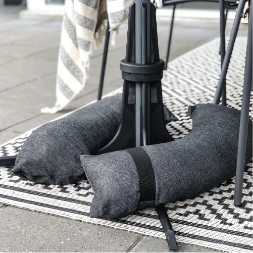 Baser Sonnenschirmständer mit zwei grauen befüllten Sandsäcken. Der Sonnenschirmständer steht vor einem Tisch auf einem schwartz-weiß karierten Teppich, der auf hellgrauen Fliesen liegt.