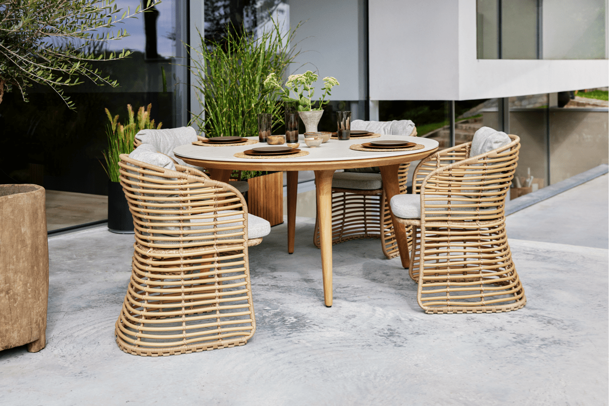 Modern outdoor teak basket chairs with light cushions around a round teak table with white stone top.