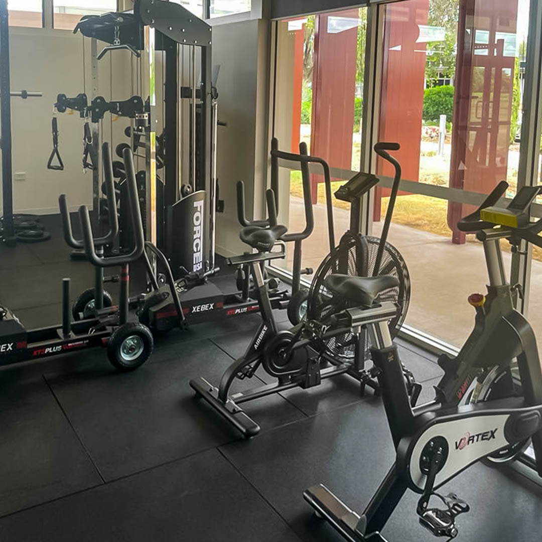 Stationary bikes and a sled lined up side by side in a high school gym fit-out, offering both cardio and strength training options for students.