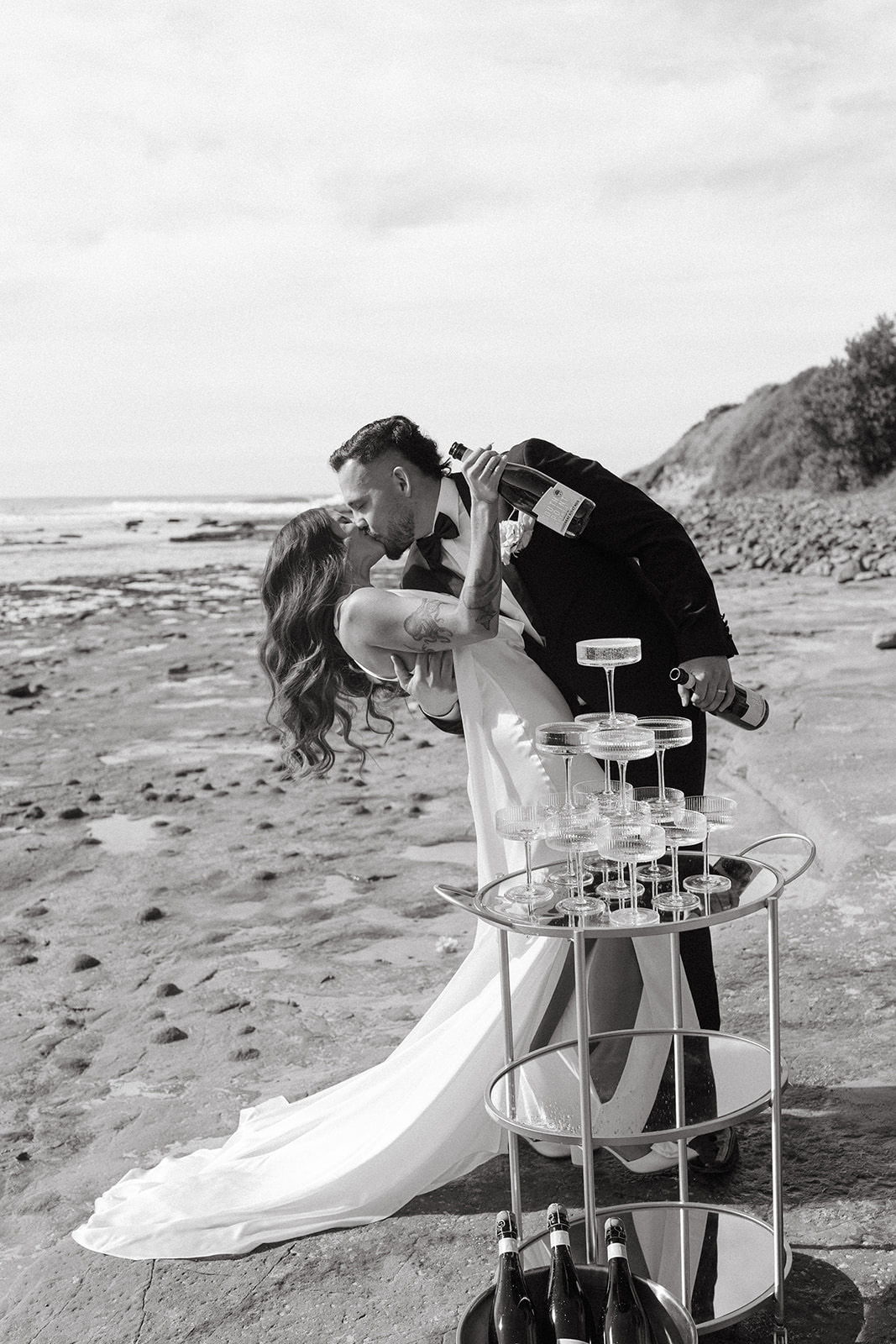 Couple kissing on the beach with champagne and wedding attire.
