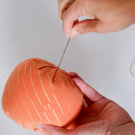 Hand holding a big needle threaded with embroidery floss ready to pull it through the stuffed fabric pumpkin