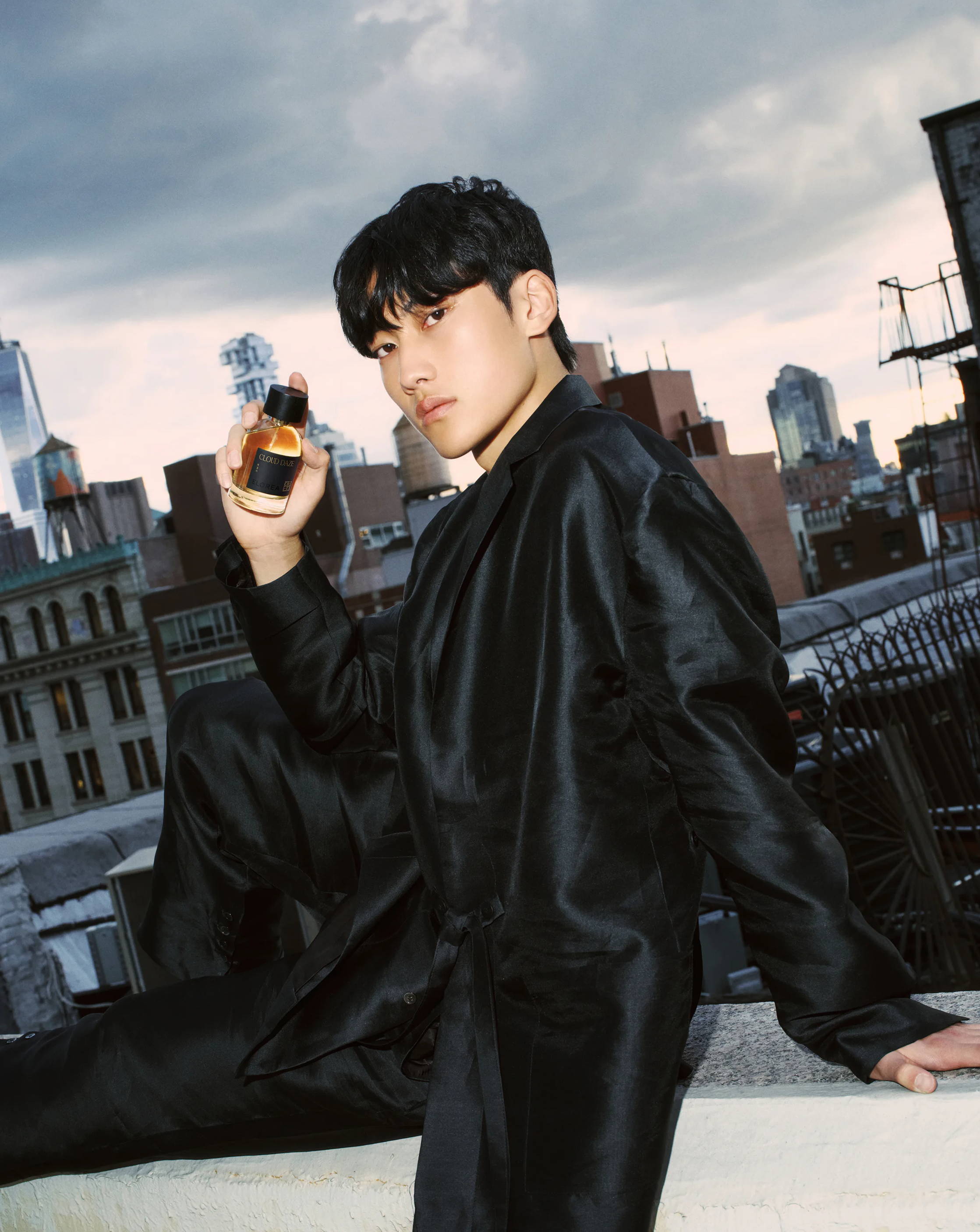 Person in black attire holding a bottle, sitting on a rooftop with city view.