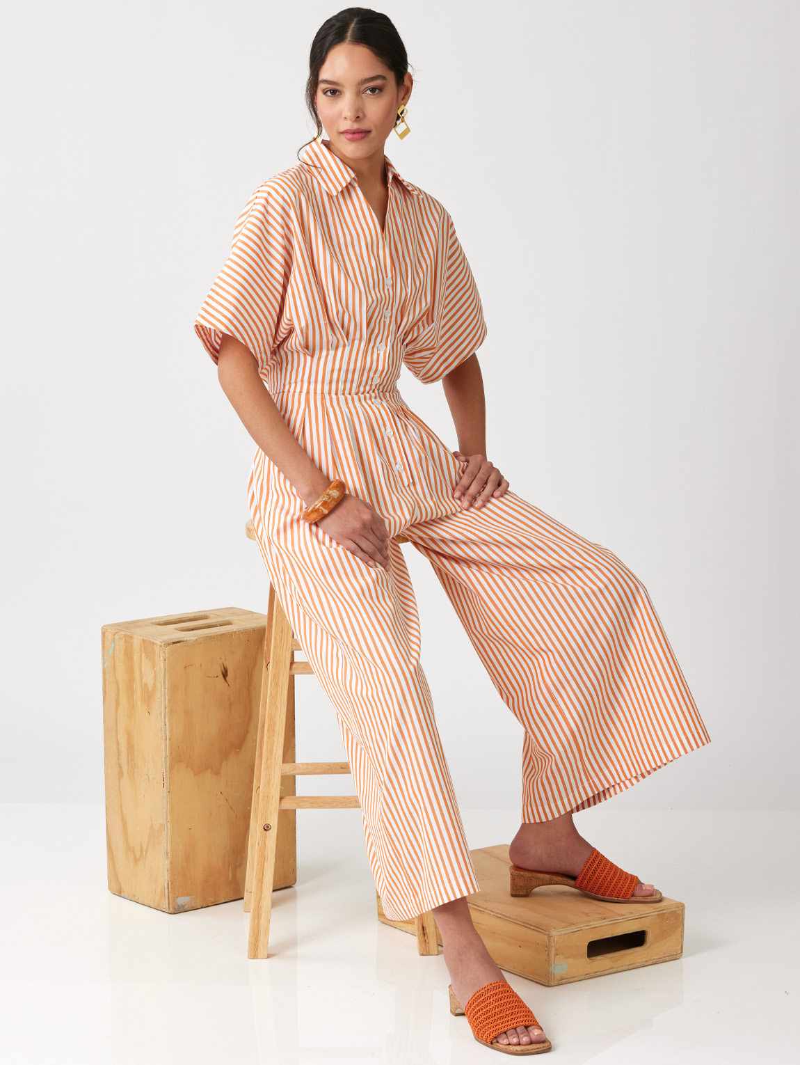 Woman in striped jumpsuit sitting on a stool, wearing orange shoes.