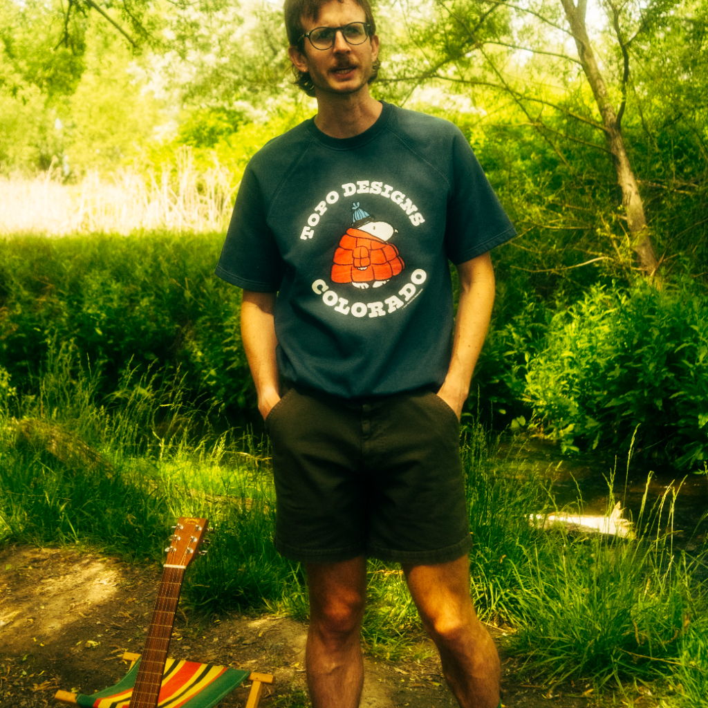 Person standing in nature, wearing a t-shirt and shorts, with a guitar nearby.