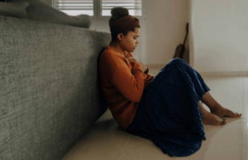 Person sitting against a wall, looking at their phone, wearing casual clothing.