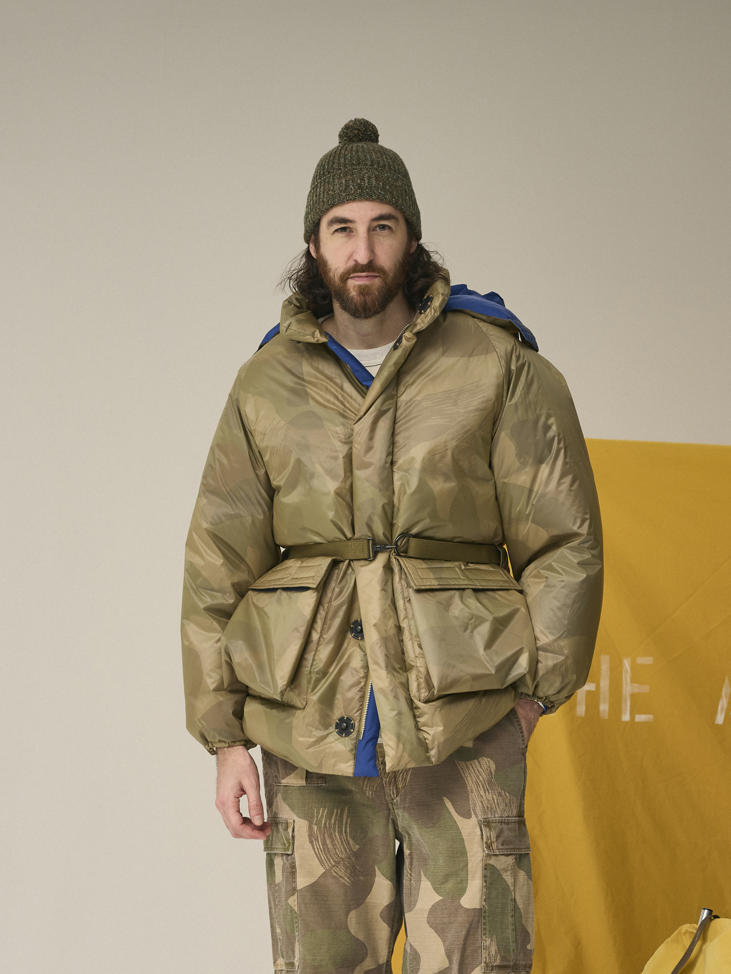 A man in a winter coat stands next to a yellow banner and backpack.