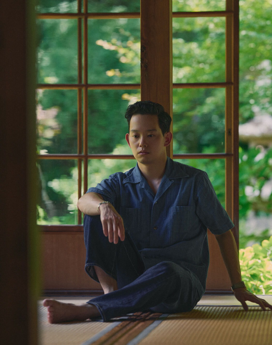 Person sitting on the floor in a room with large windows and greenery outside.