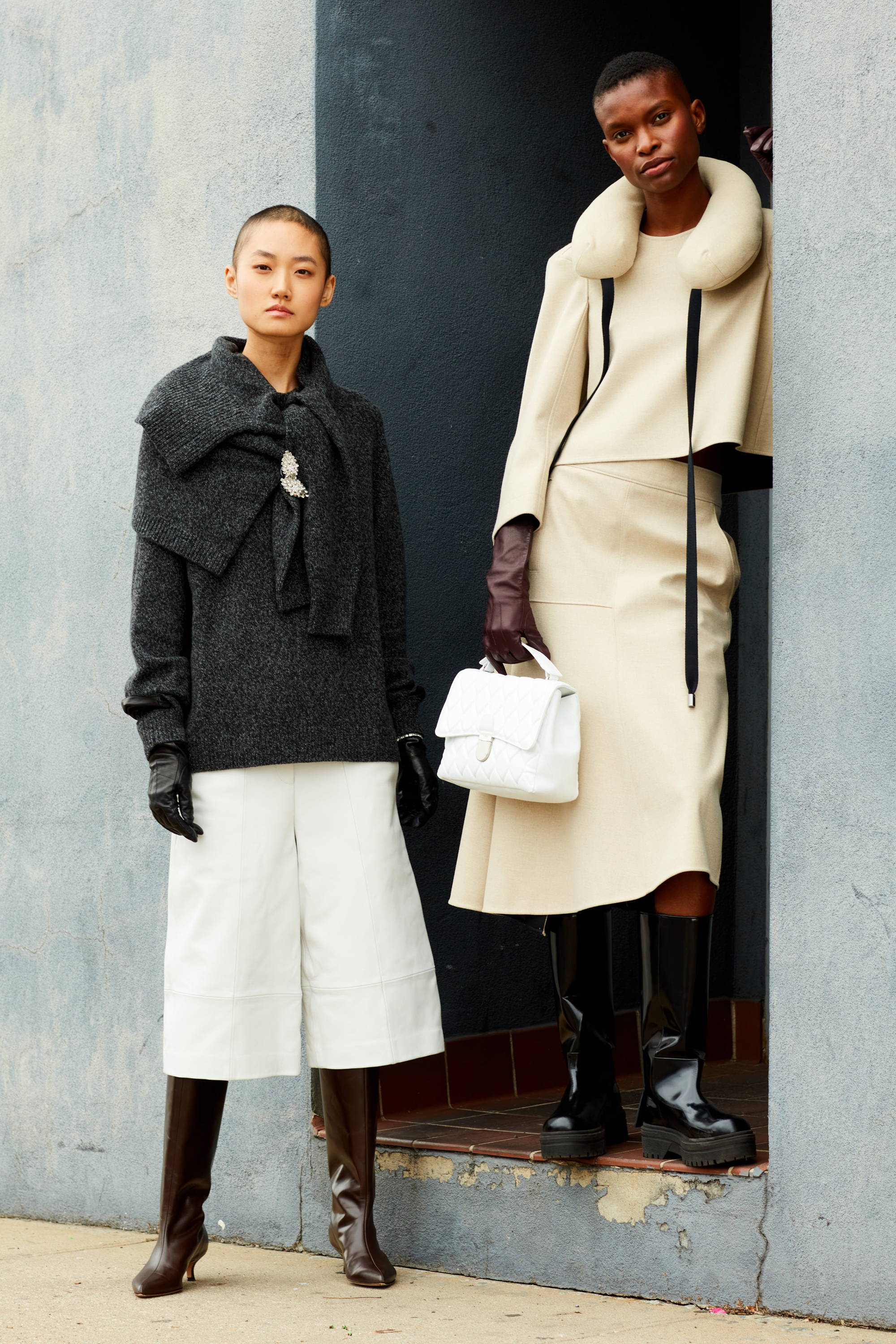 Two models in stylish outfits posed against a textured wall.