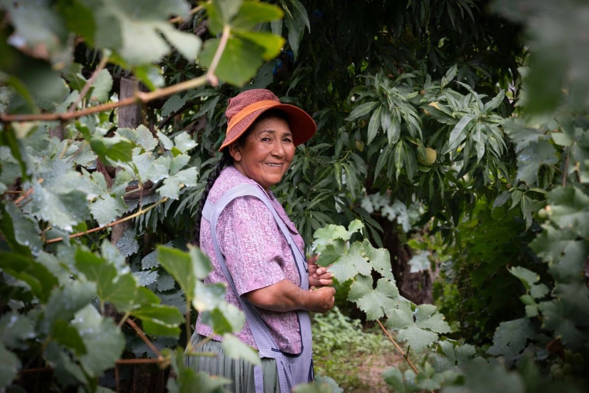 Smiling woman in a hat standing among green foliage.